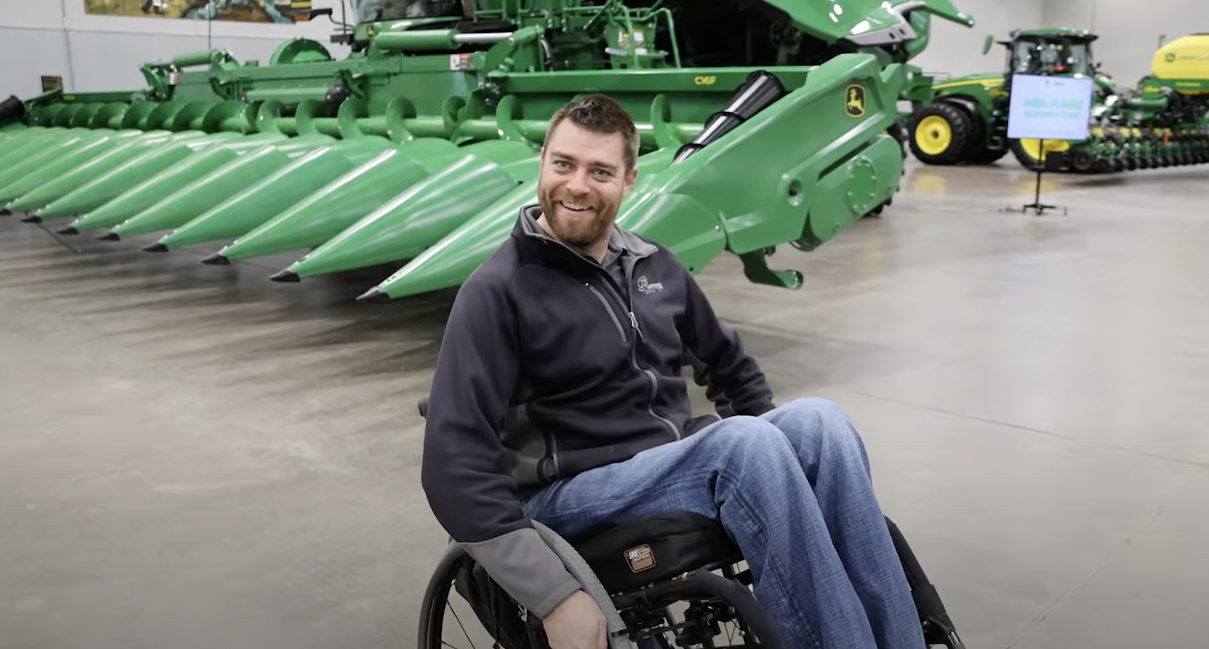 Bud Kern, doing a wheelie in his wheelchair and smiling in front of a row of John Deere machines.